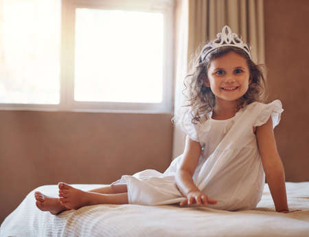 Is she not just the cutest princess youve ever seen. Portrait of an adorable little girl wearing a tiara and sitting on a bed at home.の写真素材