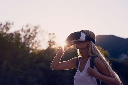 Woman hiking at sunrise using VR goggles to explore the metaverse. Blond woman on holiday hiking in a forest using VR goggles to experience augmented virtual realityの写真素材