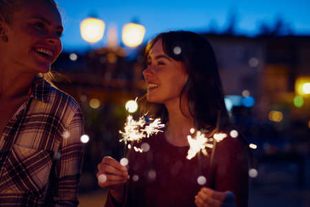 Young woman smiling at her friend and celebrating playing with fireworks. Two friends on holiday together celebrating with fireworks at night.Happy woman on holiday with friend playing with fireworksの写真素材
