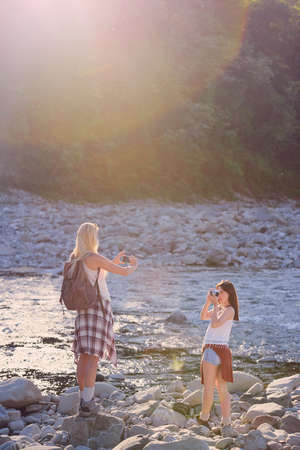 Young women taking photos of each other with a smartphone and digital camera next to a river during a hike.Two friends taking photos of each other with a camera and cellphone on a hike.の写真素材