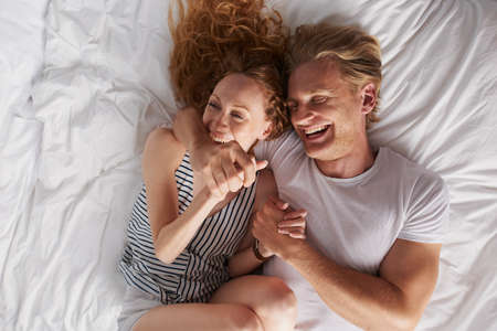 A beautiful woman playfully biting the arm of her boyfriend while they lie on their bedの写真素材