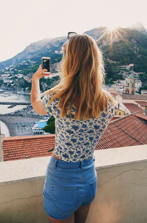 Woman taking photos of Italy coast using smartphone. Rear view of woman taking pictures of ocean view on vacationの写真素材