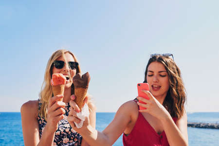 Two friends taking photos of ice cream eating on beach using smart phone technology on adventure travelの写真素材