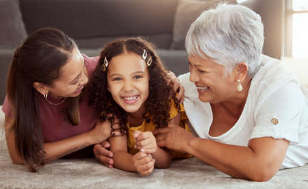 Portrait of mixed race child with single mother and grandmother in home living room. Smiling hispanic girl bonding with parent and senior in lounge. Happy three generations lying together on floorの写真素材