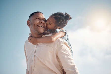 Smiling mixed race single father carrying little daughter on piggyback with copyspace. Adorable, happy, hispanic girl bonding with parent and kissing cheek on beach. Man and child enjoying free timeの写真素材