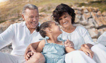 Smiling mixed race grandparents sitting with granddaughter on a beach. Adorable, happy, hispanic...