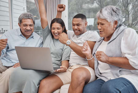 Happy and affectionate mixed race family of four using a laptop to watch sports and cheer on their favourite team in the home living room. Married couple sitting with their mature parents on the sofaの写真素材