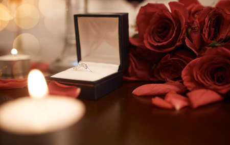 Closeup of romantic engagement ring box, fresh red roses and candles in an empty room at night....