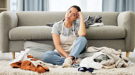 A young mixed race domestic cleaner looking distracted and overwhelmed while folding laundry. A...