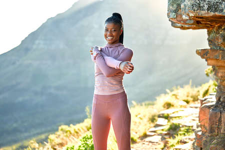 Smiling young african american woman hiking on a trail, stretching her arms and shoulder. Portrait of a smiling young athletic woman doing a warmup before hiking in nature on a mountain in the morningの写真素材