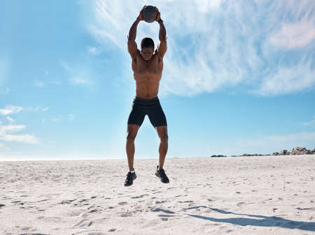 A handsome young african american male athlete working out on the beach. Dedicated black man exercising with a medicine ball outside on the sand. Committed to a healthy lifestyle and getting fitの写真素材