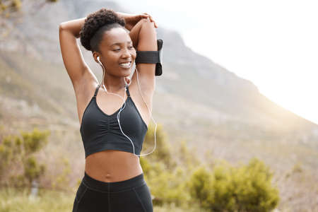 One african american female athlete with an afro listening to music on her earphones while exercising outdoors in nature. Dedicated black woman smiling while warming up before a workout outsideの写真素材