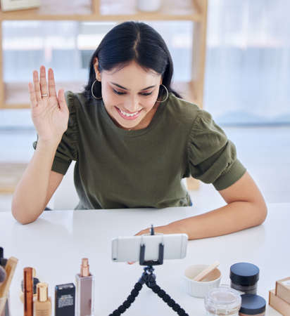 Hispanic beauty blogger waving at her smart phone while recording a video or broadcasting live. Beautiful young influencer sharing her beauty products and regimeの写真素材