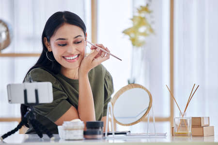Young beauty blogger smiling while looking into a vanity mirror and doing a make-up tutorial while recording with her smart phone. Influencer sharing her beauty products and tips on social networkの写真素材