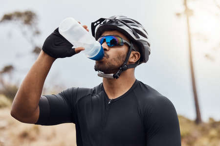Thirsty cyclist taking a break and drinking water from a bottle. Fit young man wearing glasses and a helmet while drinking water and standing outside. Athletic man cycling in nature environmentの写真素材