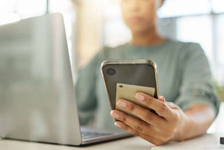 Closeup of a mixed race woman holding a phone in her hand while working on a laptop at home. One...