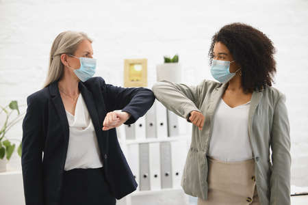 Two diverse colleagues talking and greeting each other with their elbows while wearing masks to...