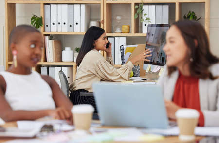 Serious ambitious indian business woman using cellphone to talk to clients while using office computer. Confident ethnic professional sitting behind team, explaining and sharing ideas while networkingの写真素材