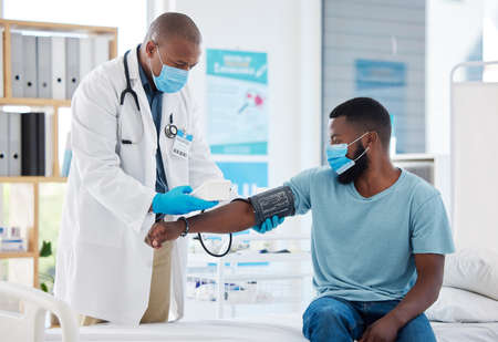 Doctor checking the blood pressure of a patient using a cuff and monitor. African american doctor reading a patients blood pressure. Medical gp and patient wearing masks to protect from covidの写真素材
