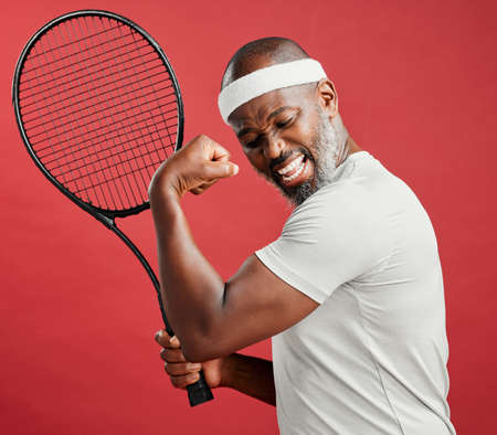 One happy mature african american man standing against a red background in studio, flexing his bicep while posing with a tennis racquet. Smiling black man feeling fit and sporty while playing a matchの写真素材