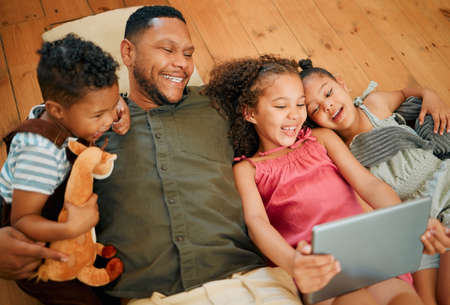 A happy mixed race family of four relaxing and lying on the lounge floor together. Loving black single parent bonding with his kids while using a digital tablet to watch moviesの写真素材