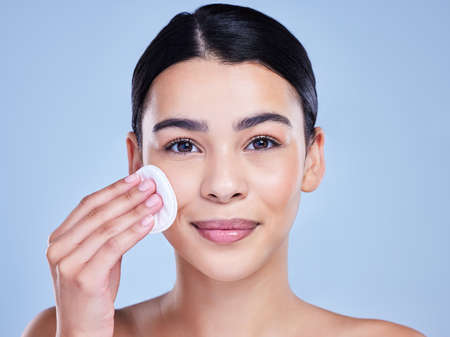 Studio Portrait of a beautiful mixed race woman using a cotton pad to remove makeup during a selfcare grooming routine. Hispanic woman applying cleanser to her face against blue copyspace backgroundの写真素材