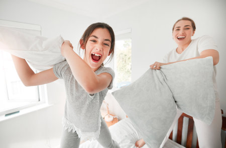 Portrait of cute teenage daughter and mother having a fun pillow fight at home. Happy young girl and mother holding pillows and standing on bed playing and enjoying free time together in the morningの写真素材