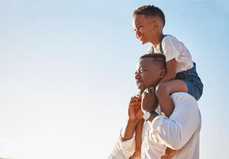 Young african american father carrying his son on his shoulders against a blue sky while walking outdoors on a sunny day. Excited little boy having fun with his dad and enjoying summer vacationの写真素材