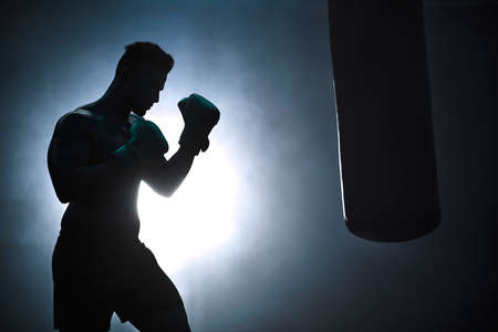 Silhouette of an unrecognizable boxer man sparring with a punching bag in a dark gym. Male athlete working out in preparation for his upcoming fight. Young martial artist training and practicingの写真素材