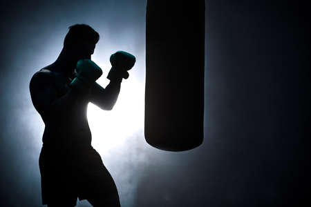 Silhouette of an unrecognizable boxer man sparring with a punching bag in a dark gym. Male athlete working out in preparation for his upcoming fight. Young martial artist training and practicingの写真素材