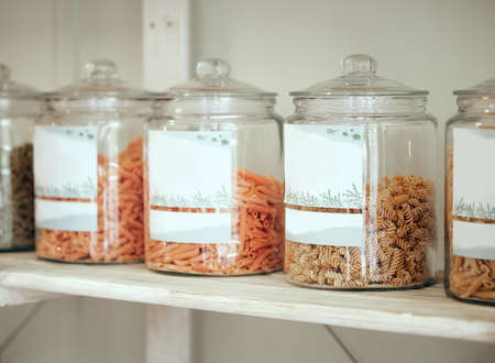 Glass jars of different types of pasta lined up in a row on a display shelf in a grocery store. Get...