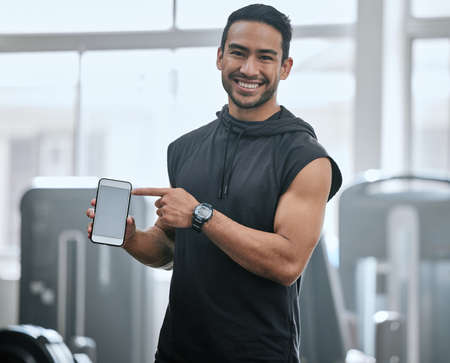 Portrait of smiling asian trainer alone in gym showing cellphone screen. Coach standing, promoting discount and deal for workout in health club. Young confident man tracking exercise in fitness centreの写真素材