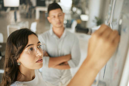 Focused business woman coach mentor writing on board planning strategy while brainstorming with colleagues. Diverse colleagues discussing ideas while collaborating on projectの写真素材