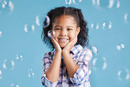 Portrait of happy smiling little girl posing with her hands on her face with bubbles falling around her against blue studio background. Cute mixed race kid in casual clothesの写真素材