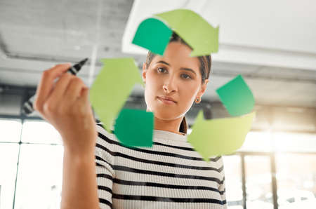 Young focused mixed race businesswoman drawing a recycle symbol on a glass window in an office at...