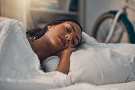 A beautiful young mixed race woman sleeping in a soft comfortable bed at home. One hispanic female feeling exhausted and resting in a comforting bedの写真素材