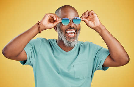 Happy mature African American man standing alone against yellow background in a studio and posing with sunglasses. Smiling black man feeling fashionable and cool while wearing glasses. Summer and beach.の写真素材