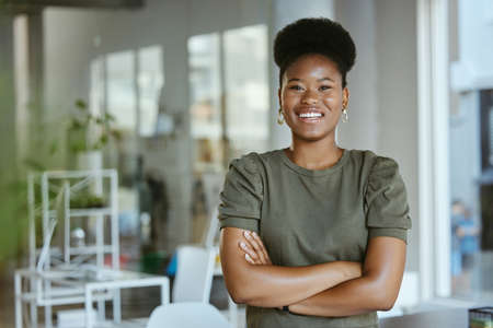 Young happy african american businesswoman standing with her arms crossed alone at work. One cheerful black female boss with an afro smiling while standing in an officeの写真素材
