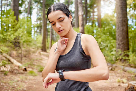 One beautiful young fit and athletic woman checking her pulse while exercising outdoors. An attractive mixed race female checking her heart rate on her smart watch during a workout in natureの写真素材