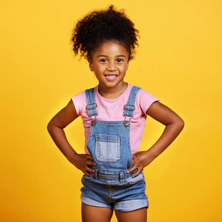 Studio portrait mixed race girl looking standing with her hands on her hips isolated against a yellow background. Cute hispanic child posing inside. Happy and cute kid smiling and looking confidentの写真素材