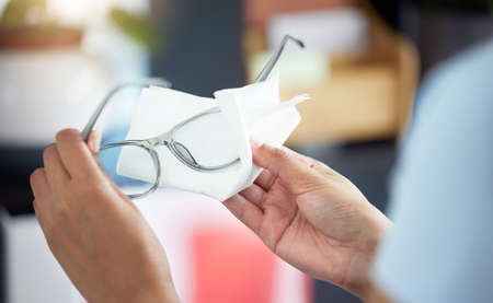 An unrecognizable woman cleaning her glasses in her apartment. One unknown woman using a tissue to remove dust from her spectaclesの写真素材