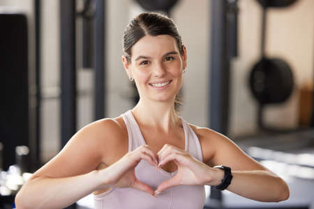 Portrait of smiling caucasian athlete showing heart shape sign and symbol during workout in gym. Strong, fit, active woman showing passion and love for exercise and training in health and sports clubの写真素材