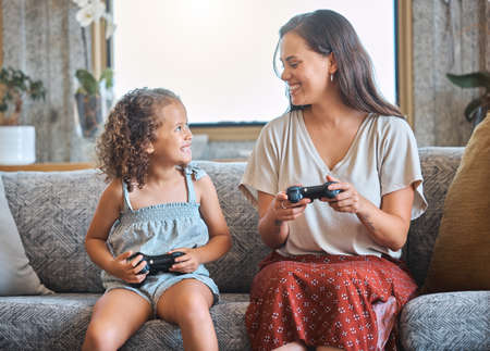 Hispanic mother and daughter playing video games together while sitting on the couch at home. Fun young mother and daughter using joysticks while playing and spending free time together on weekendの写真素材