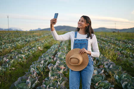 Woman farmer taking a selfie on her smartphone while standing in a cabbage field. Young brunette...
