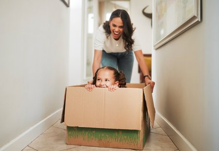 Mother and daughter playing with a cardboard box. Excited little girl sitting in a box. Parent pushing her daughter in a box. Parent having fun with her child at home. Cheerful mother and daughterの写真素材