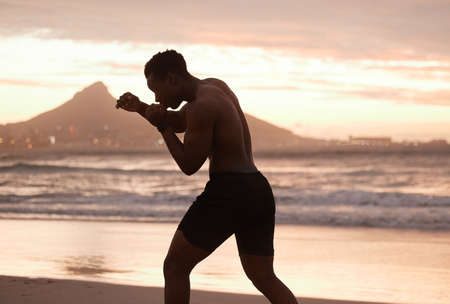 One african american man practicing shadow boxing on a beach at sunset. Black male focus on speed, strength and fitness while showing his muscular athletic shape against a sunrise copyspace backgroundの写真素材