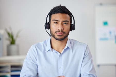 Portrait of a young mixed race businessman working in a call center. Operator at his office job. Hispanic man working, wearing a headset and working in customer service, on a call.の写真素材