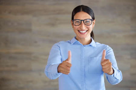 Smiling businesswoman showing thumbs up sign with copyspace. Mixed race professional standing alone and using hand gestures to symbol good luck. Hispanic woman wearing glasses endorsing and supportingの写真素材