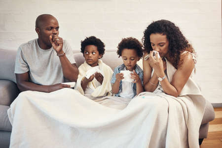 African american family of four feeling sick and unwell with flu and cold while covered in a blanket on a couch. Parents and two kids coughing and blowing nose from seasonal allergy symptoms and covidの写真素材