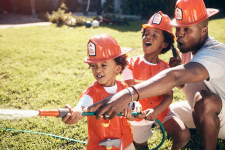 Father playing with his sons outside. Little boys dressed as firemen. African American boys playing outside. Brothers playing with a hosepipe in the garden. Siblings spraying water from a hosepipe.の写真素材